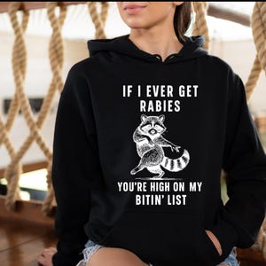 Op de afbeelding: Zwarte hoodie met een witte illustratie van een wasbeer en de tekst "IF I EVER GET RABIES YOU'RE HIGH ON MY BITIN' LIST". De wasbeer is afgebeeld in een speelse pose. De hoodie heeft een capuchon met trekkoord en een voorzak.