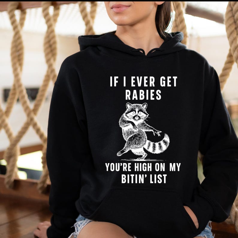 Op de afbeelding: Zwarte hoodie met een witte illustratie van een wasbeer en de tekst "IF I EVER GET RABIES YOU'RE HIGH ON MY BITIN' LIST". De wasbeer is afgebeeld in een speelse pose. De hoodie heeft een capuchon met trekkoord en een voorzak.