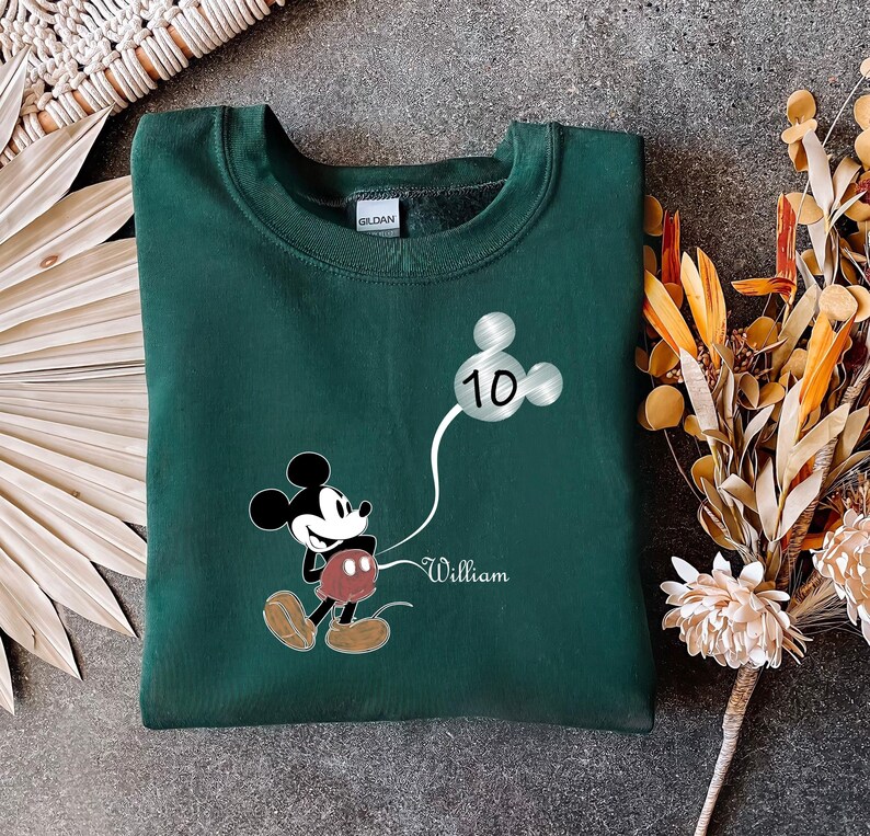 Puede incluir: Sudadera verde con un gr&aacute;fico de Mickey Mouse sosteniendo un globo plateado con el n&uacute;mero 10. El texto "William" est&aacute; impreso debajo del gr&aacute;fico.