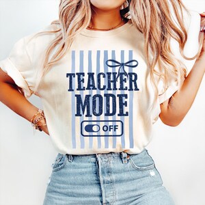 May include: Cream-colored t-shirt with the text "TEACHER MODE" in navy blue, set against a backdrop of light blue vertical stripes. Below the text is an "OFF" switch graphic. A navy blue bow is above the text.