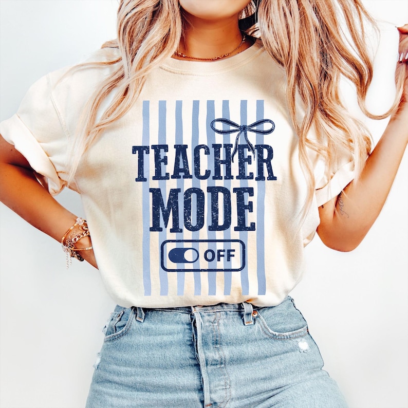 May include: Cream-colored t-shirt with the text "TEACHER MODE" in navy blue, set against a backdrop of light blue vertical stripes. Below the text is an "OFF" switch graphic. A navy blue bow is above the text.