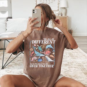May include: Brown t-shirt with a colorful graphic of various sea creatures, including a whale, octopus, and fish. The text "We Are All DIFFERENT But In This School We All SWIM TOGETHER" is displayed in a retro font.