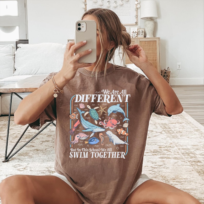 May include: Brown t-shirt with a colorful graphic of various sea creatures, including a whale, octopus, and fish. The text "We Are All DIFFERENT But In This School We All SWIM TOGETHER" is displayed in a retro font.
