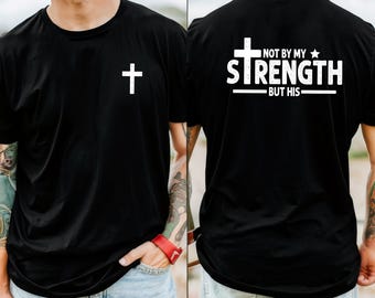 May include: Black t-shirt with a white cross on the front. The back of the shirt features the text "NOT BY MY STRENGTH BUT HIS" in white, with a cross and star graphic. The shirt is a crew neck style.