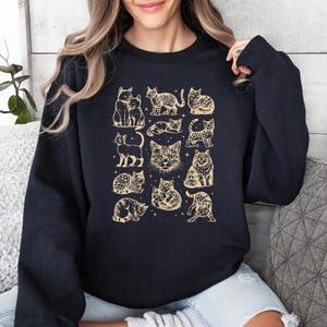 May include: Navy blue sweatshirt featuring a repeating pattern of various cat illustrations in a vintage style. The cats are depicted in different poses and are in a light tan color. The sweatshirt is a crew neck style.