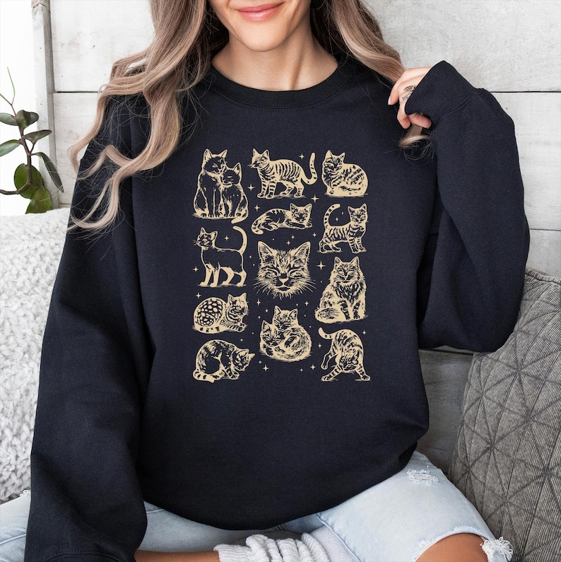 May include: Navy blue sweatshirt featuring a repeating pattern of various cat illustrations in a vintage style. The cats are depicted in different poses and are in a light tan color. The sweatshirt is a crew neck style.
