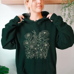 May include: Dark green hoodie featuring a floral design. The hoodie has a drawstring hood and a kangaroo pocket. The floral design is a detailed line drawing of various flowers and foliage in a light beige color. The hoodie is made of a soft, comfortable material.