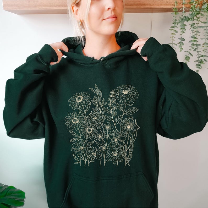 May include: Dark green hoodie featuring a floral design. The hoodie has a drawstring hood and a kangaroo pocket. The floral design is a detailed line drawing of various flowers and foliage in a light beige color. The hoodie is made of a soft, comfortable material.