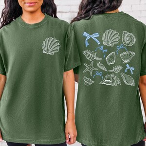 Puede incluir: Camiseta verde oliva con un dise&ntilde;o de concha blanca en la parte delantera y una colecci&oacute;n de conchas marinas y lazos azules en la parte trasera. La camiseta tiene un corte holgado y mangas cortas.