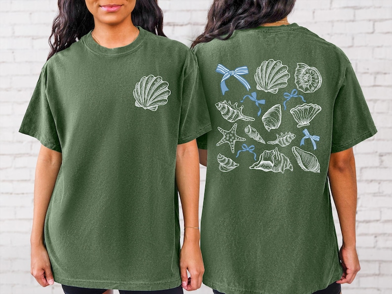 Puede incluir: Camiseta verde oliva con un dise&ntilde;o de concha blanca en la parte delantera y una colecci&oacute;n de conchas marinas y lazos azules en la parte trasera. La camiseta tiene un corte holgado y mangas cortas.