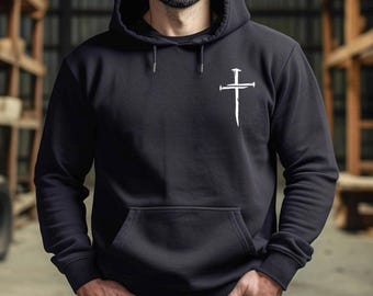 May include: Black hooded sweatshirt with a white cross graphic on the chest. The sweatshirt has a front pocket and a drawstring hood. The background is a blurred warehouse setting.