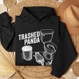 May include: Black hoodie with a white graphic reading "Trashed Panda". The design includes a bin, a toilet, and a raccoon. The hoodie has a drawstring hood and long sleeves.