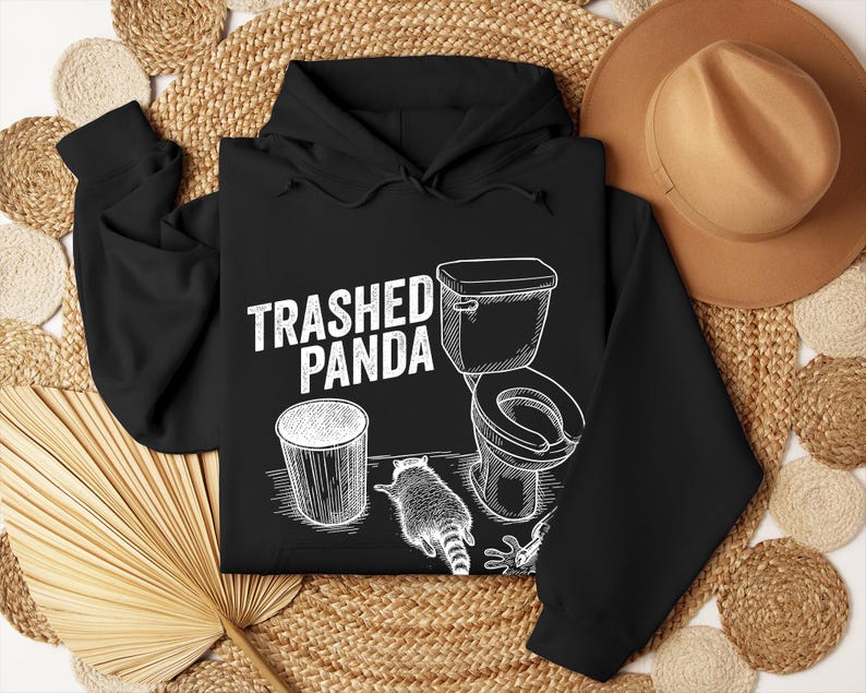May include: Black hoodie with a white graphic reading "Trashed Panda". The design includes a bin, a toilet, and a raccoon. The hoodie has a drawstring hood and long sleeves.