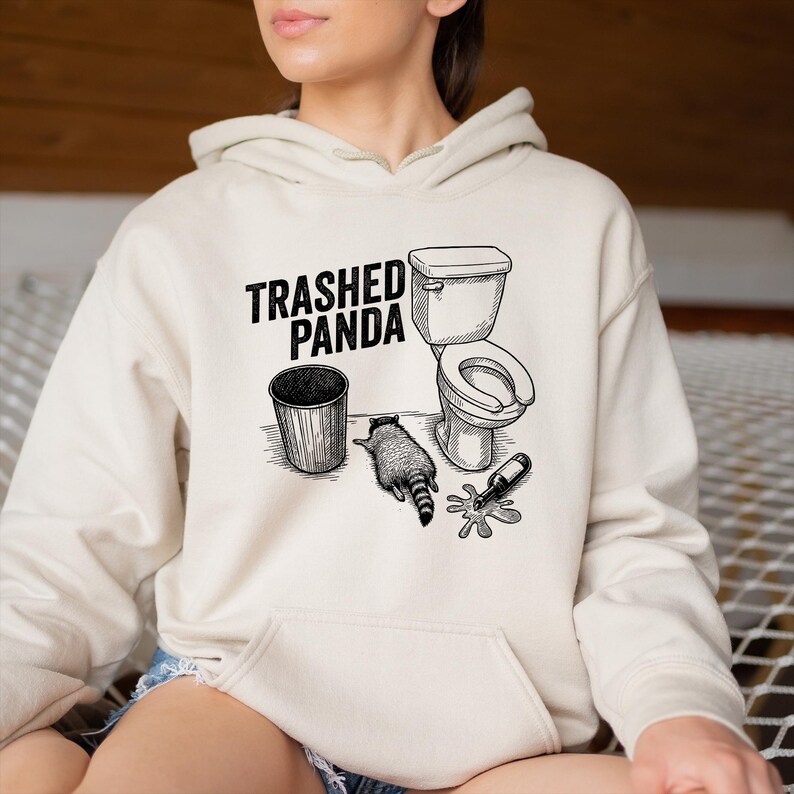 May include: Cream-coloured hoodie with a black and white graphic. The graphic features the text "Trashed Panda" above a bin, a toilet, a raccoon, and a spilled bottle. The hoodie has a front pocket and a hood.