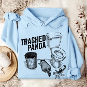 May include: Pale blue hoodie featuring the words "Trashed Panda" and a monochrome illustration of a toilet, bin, raccoon, and a spilled bottle. The design is in black and white.
