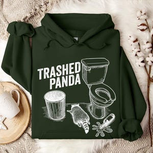 May include: Dark green hoodie featuring the text "Trashed Panda" and a white graphic of a toilet, bin, raccoon, and spilled bottle. The hoodie has a hood and a front pocket.