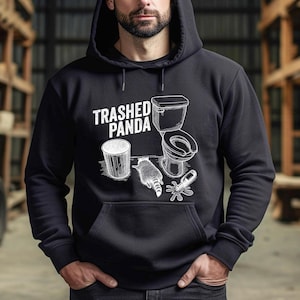 The image displays a raccoon lying face down on the floor in a comical pose. Surrounding items include a toilet, a garbage can, and a spilled glass bottle. The caption TRASHED PANDA is written in a distressed font, highlighting the drunk or hungover theme.