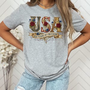 May include: A light grey t-shirt with the word "USA" in a decorative font, adorned with floral and ribbon details. The shirt also features the text "250 YEARS OF FREEDOM" on a banner. The design incorporates red, white, blue, and gold colours.