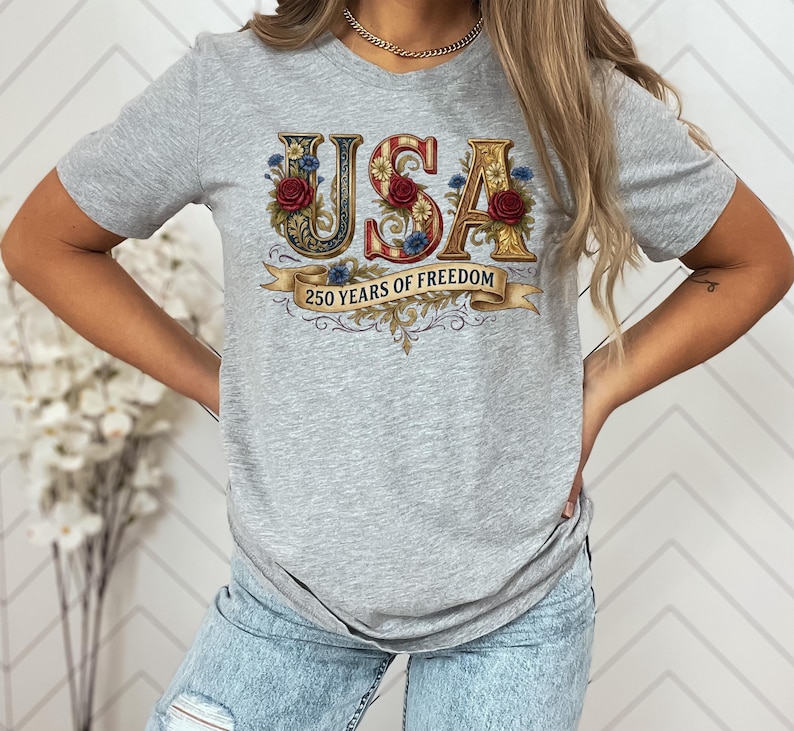May include: A light grey t-shirt with the word "USA" in a decorative font, adorned with floral and ribbon details. The shirt also features the text "250 YEARS OF FREEDOM" on a banner. The design incorporates red, white, blue, and gold colours.