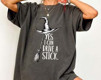 May include: Dark gray t-shirt featuring a witch hat and broom graphic with the text "YES I CAN DRIVE A STICK." The design is in black and white. The shirt has short sleeves and a relaxed fit.