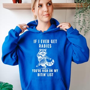 Op de afbeelding: Een koningsblauwe hoodie met een witte afbeelding van een wasbeer en de tekst "IF I EVER GET RABIES YOU'RE HIGH ON MY BITIN' LIST". De hoodie heeft een capuchon met trekkoord en een voorzak.
