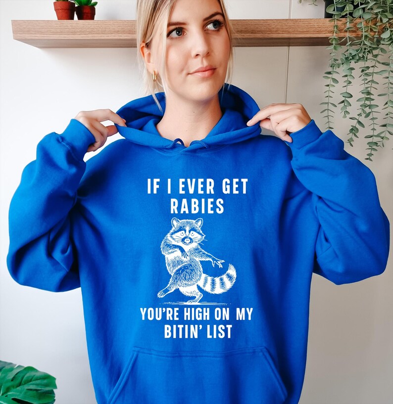 Op de afbeelding: Een koningsblauwe hoodie met een witte afbeelding van een wasbeer en de tekst "IF I EVER GET RABIES YOU'RE HIGH ON MY BITIN' LIST". De hoodie heeft een capuchon met trekkoord en een voorzak.