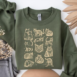 May include: Olive green sweatshirt featuring a beige illustration of various cat poses. The graphic includes cats sitting, standing, and lounging, with a few stars scattered around the images. The sweatshirt has a crew neck and long sleeves.