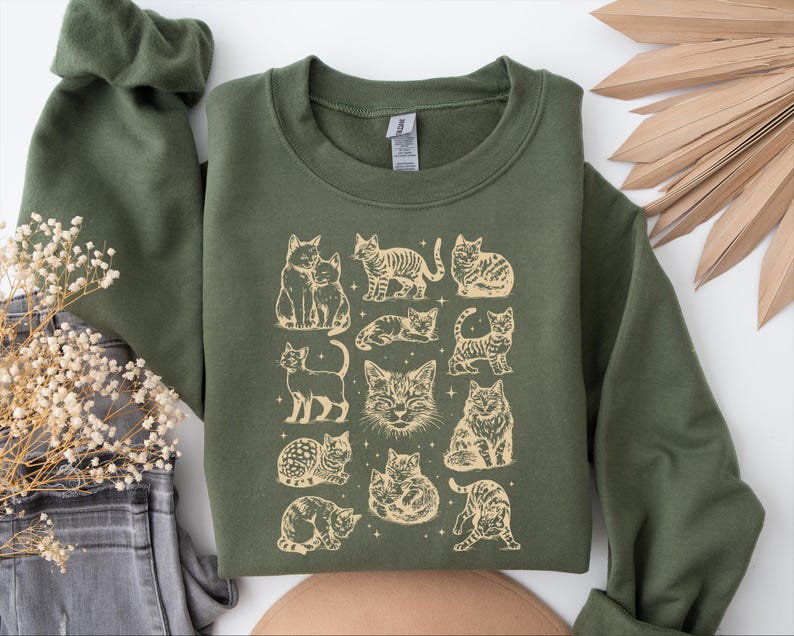 May include: Olive green sweatshirt featuring a beige illustration of various cat poses. The graphic includes cats sitting, standing, and lounging, with a few stars scattered around the images. The sweatshirt has a crew neck and long sleeves.