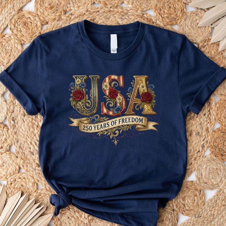 Navy blue t-shirt with the word "USA" in ornate gold lettering, accented with red roses and blue floral details. A banner below reads "250 YEARS OF FREEDOM".
