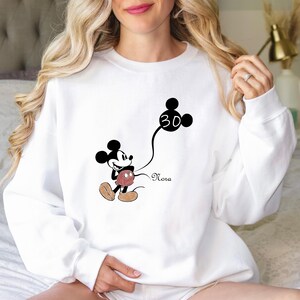 Puede incluir: Sudadera blanca con un gr&aacute;fico de Mickey Mouse en blanco y negro sosteniendo un globo con el n&uacute;mero 30. El texto "Nora" est&aacute; impreso debajo del gr&aacute;fico.