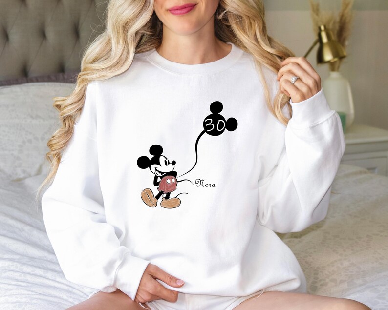 Puede incluir: Sudadera blanca con un gr&aacute;fico de Mickey Mouse en blanco y negro sosteniendo un globo con el n&uacute;mero 30. El texto "Nora" est&aacute; impreso debajo del gr&aacute;fico.