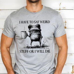 May include: A light gray t-shirt featuring a whimsical illustration of a frog holding a mug. The frog is drawn in black and white, with the text "I HAVE TO SAY WEIRD STUFF OR I WILL DIE" printed above and below the frog.