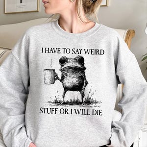 May include: A light gray sweatshirt featuring a black and white illustration of a frog holding a mug. The text "I HAVE TO SAY WEIRD STUFF OR I WILL DIE" is printed above and below the frog graphic. The sweatshirt is made of a soft material.