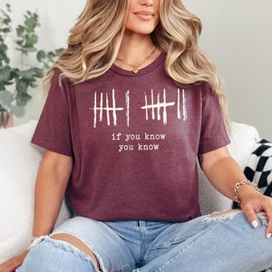 May include: Burgundy t-shirt with white text and tally marks. The text reads "if you know you know". The shirt has a V-neck and short sleeves. The shirt is made of a soft material.