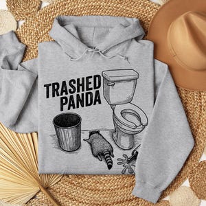 May include: A light grey hoodie featuring the text "Trashed Panda" above a graphic of a toilet, bin, and raccoon. The raccoon is lying down. The hoodie has a drawstring hood.