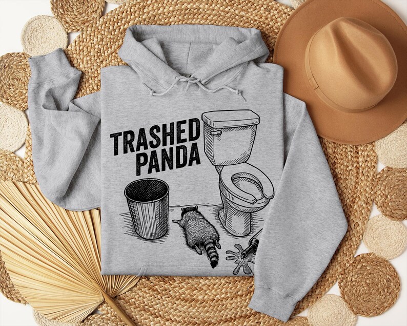 May include: A light grey hoodie featuring the text "Trashed Panda" above a graphic of a toilet, bin, and raccoon. The raccoon is lying down. The hoodie has a drawstring hood.