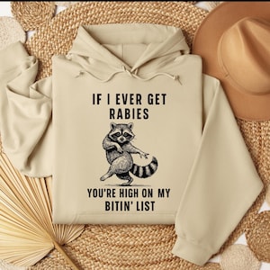 Hoodie with a funny raccoon and the phrase “If I Ever Get Rabies You’re High On My Bitin’ List.” Trendy viral meme design with sarcastic humor.