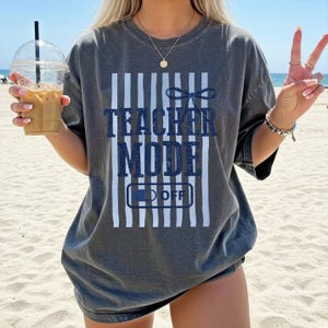 May include: Dark gray t-shirt with the text "TEACHER MODE OFF" in navy blue. The design includes vertical white stripes and a bow graphic. The shirt is a casual, oversized style, suitable for everyday wear.