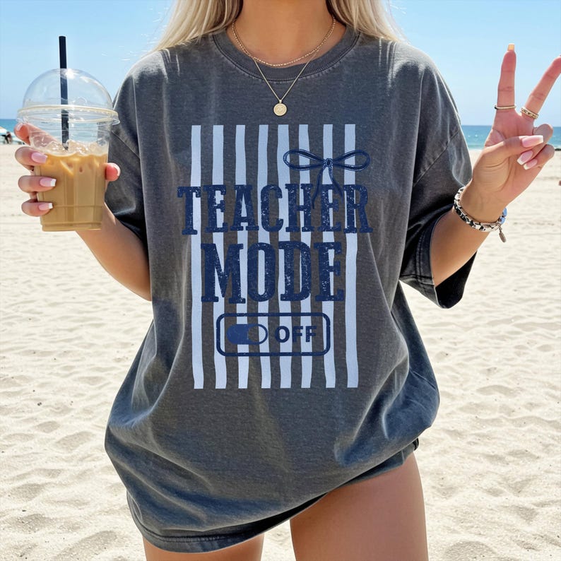May include: Dark gray t-shirt with the text "TEACHER MODE OFF" in navy blue. The design includes vertical white stripes and a bow graphic. The shirt is a casual, oversized style, suitable for everyday wear.