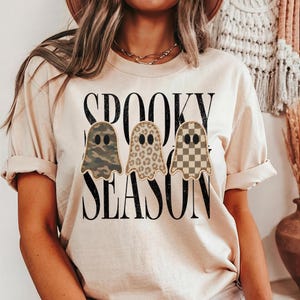 May include: A beige short-sleeve t-shirt featuring the black text SPOOKY SEASON. Below the text are three ghost illustrations with different patterns: one with camouflage, one with leopard print, and one with a tan and brown checkerboard pattern.