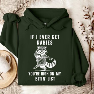 Op de afbeelding: Donkergroene hoodie met een witte wasbeer-afbeelding en de tekst "IF I EVER GET RABIES YOU'RE HIGH ON MY BITIN' LIST". De hoodie heeft een capuchon en lange mouwen. De wasbeer staat op zijn achterpoten.