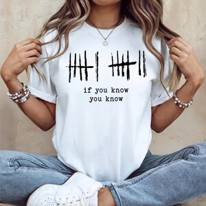 May include: White t-shirt with black vertical lines and the text "if you know you know". The shirt has short sleeves and a crew neck. The graphic is centered on the front of the shirt.