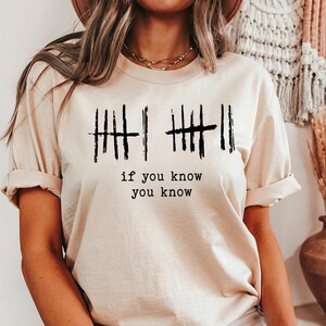 May include: Beige t-shirt with black tally marks and the text "if you know you know". The short-sleeved shirt has rolled-up sleeves and a crew neck. The shirt is made of a soft, comfortable material.
