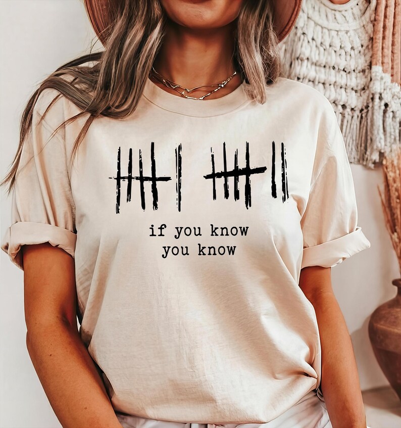 May include: Beige t-shirt with black tally marks and the text "if you know you know". The short-sleeved shirt has rolled-up sleeves and a crew neck. The shirt is made of a soft, comfortable material.