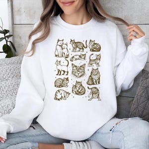 May include: White sweatshirt featuring a collection of cat illustrations in various poses and patterns. The cats are depicted in a sepia tone, creating a vintage aesthetic. The design is centered on the front of the sweatshirt.