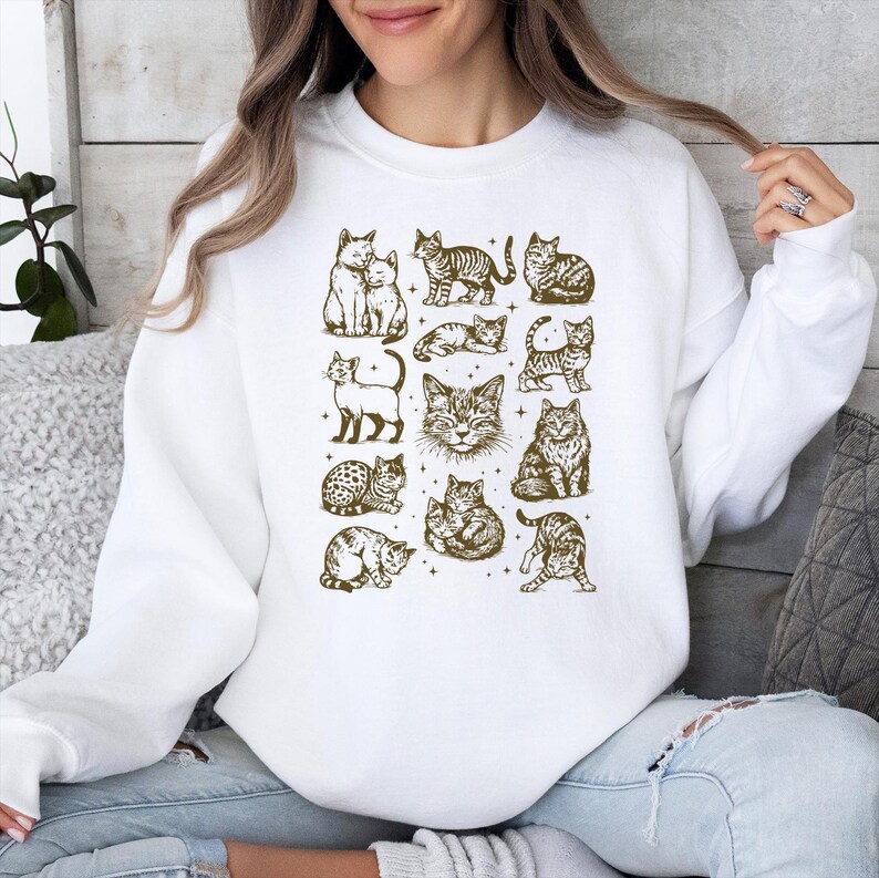 May include: White sweatshirt featuring a collection of cat illustrations in various poses and patterns. The cats are depicted in a sepia tone, creating a vintage aesthetic. The design is centered on the front of the sweatshirt.
