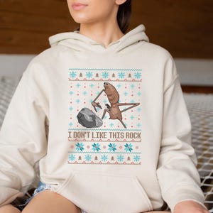 May include: Cream-colored hooded sweatshirt with a graphic of a creature and a rock, with the text "I DON'T LIKE THIS ROCK". The design includes snowflakes and trees in blue and red.