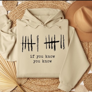 May include: A beige hoodie with a drawstring hood and a kangaroo pocket. The front of the hoodie features black tally marks and the text "if you know you know". The hoodie is made of a soft, comfortable material.