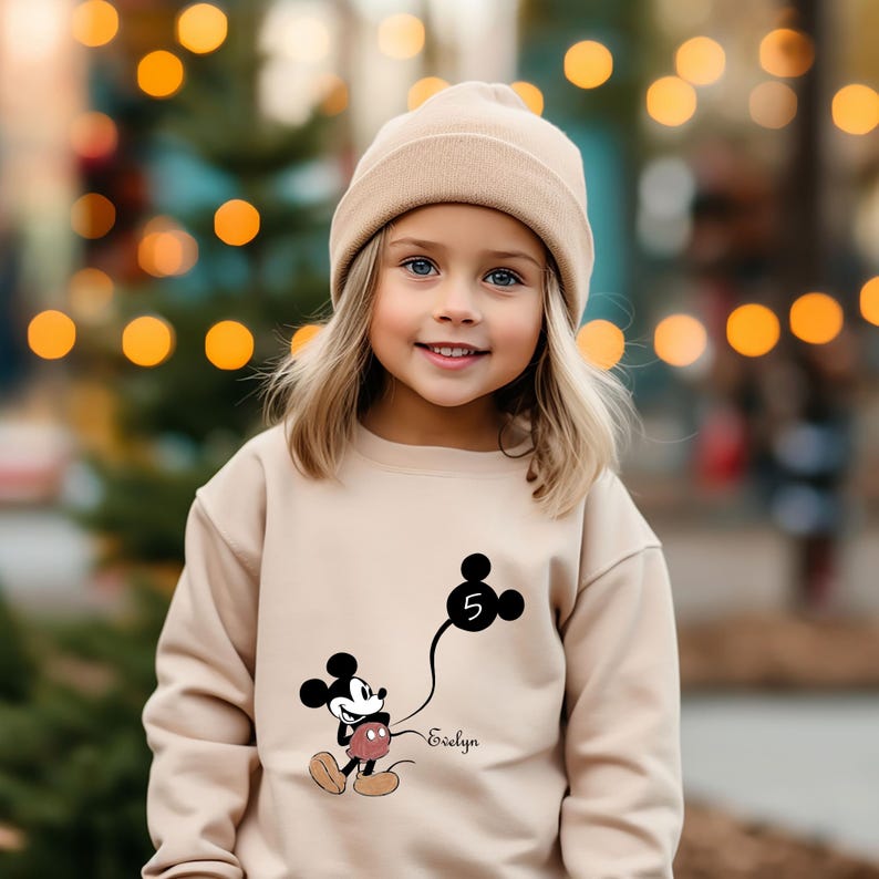 Puede incluir: Una ni&ntilde;a peque&ntilde;a con un su&eacute;ter beige con un gr&aacute;fico de Mickey Mouse negro sosteniendo un globo con el n&uacute;mero 5. El su&eacute;ter tambi&eacute;n tiene el nombre "Evelyn" impreso en &eacute;l. La ni&ntilde;a lleva una boina beige y tiene el pelo rubio.