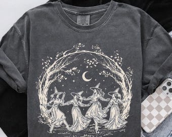 May include: Dark gray sweatshirt featuring a cream-colored illustration of four witches dancing in a circle under a crescent moon and stars. The design is framed by a tree branch archway. The sweatshirt has a crew neck and long sleeves.